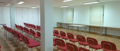 Un salón de conferencias con hileras de sillas rojas y una mesa larga, paredes blancas y luces en el techo.
