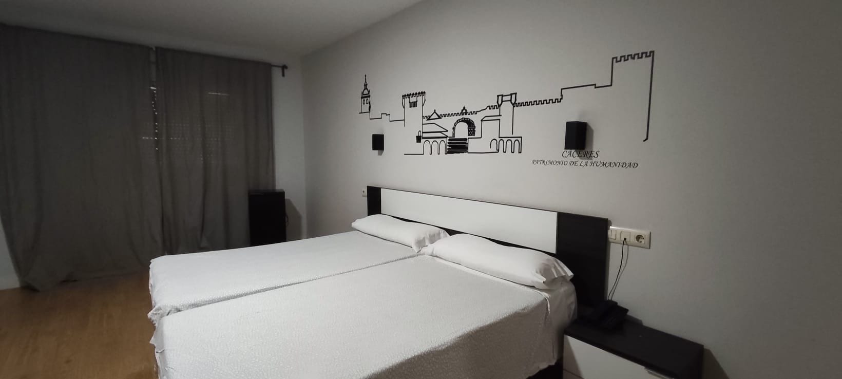 Habitación de hotel con cama, una obra de arte en la pared que representa un paisaje urbano y una ventana con cortinas.