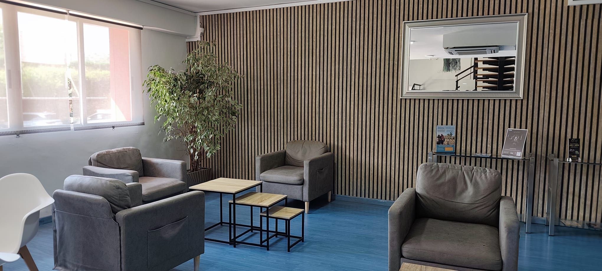 A modern waiting area with grey sofas, a small wooden table, a potted plant, and a striped wall with a mirror.