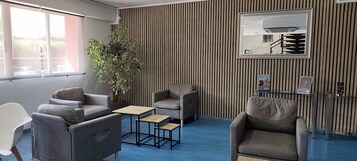 Un área de espera moderna con sofás grises, una pequeña mesa de madera, una planta en maceta y una pared a rayas con un espejo.
