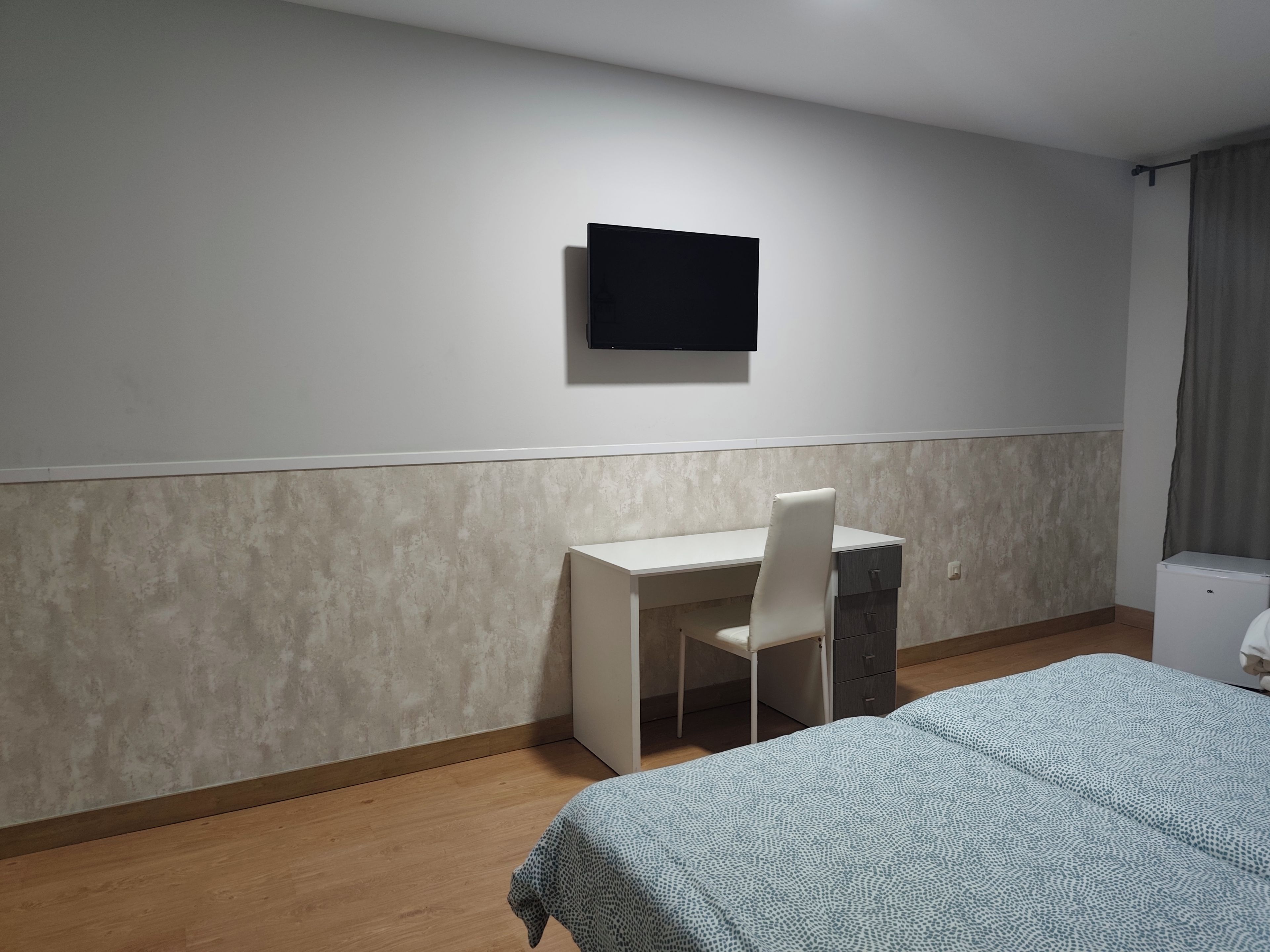 Una habitación con una cama, un escritorio, una silla, un televisor montado en la pared y un ventanal con cortinas.