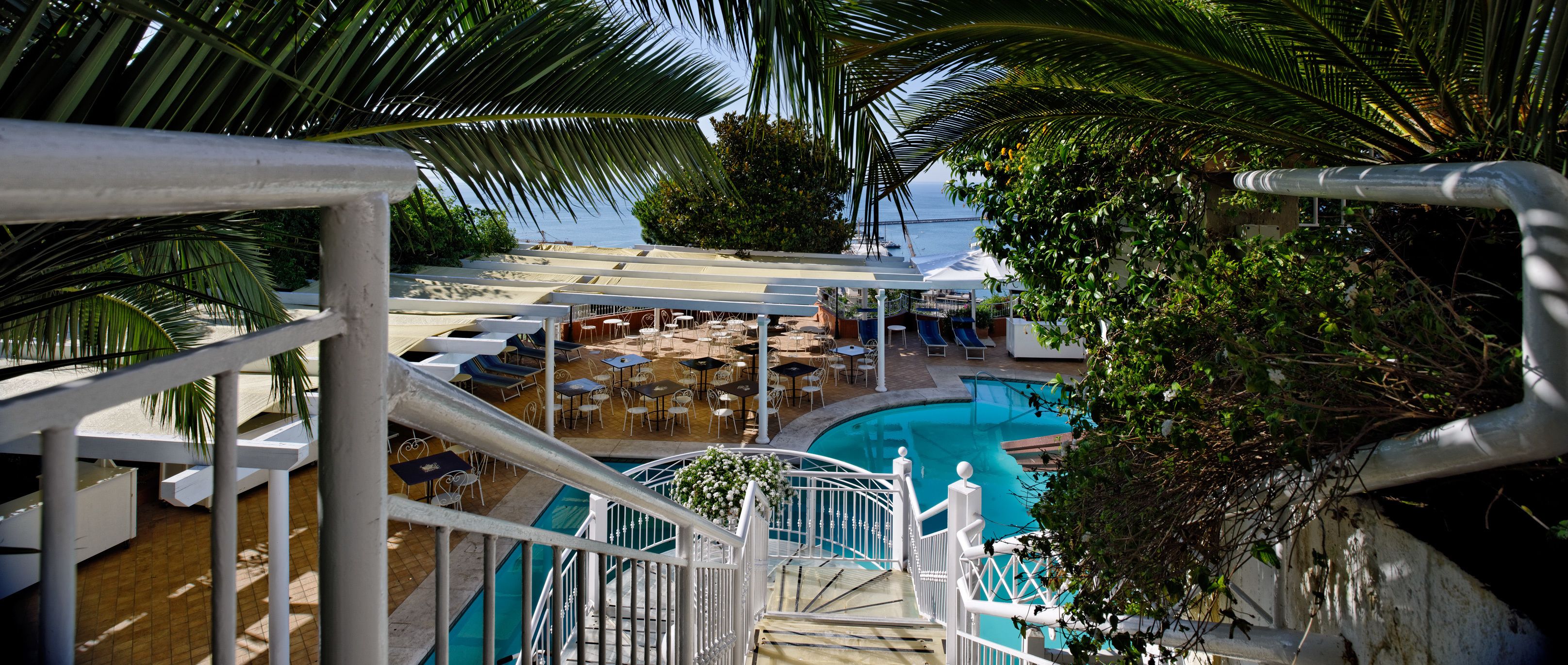 Un área de piscina con sillones de descanso y sombrillas, una escalera que lleva a una terraza, y vista al océano.