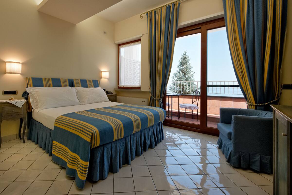 A hotel room with a bed, a nightstand, a chair, a balcony with a view, and a window with curtains.
