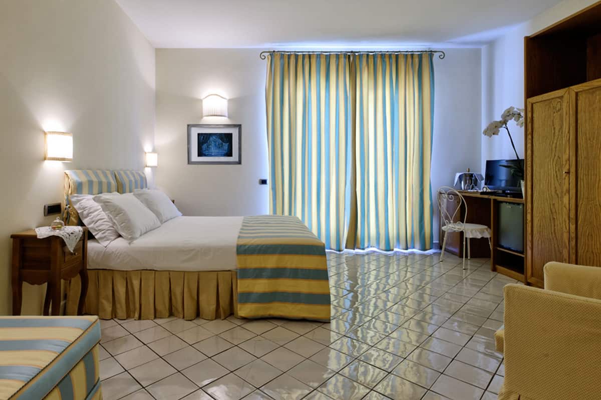 A hotel room with a bed, bedside table, lamp, and a desk with a computer.