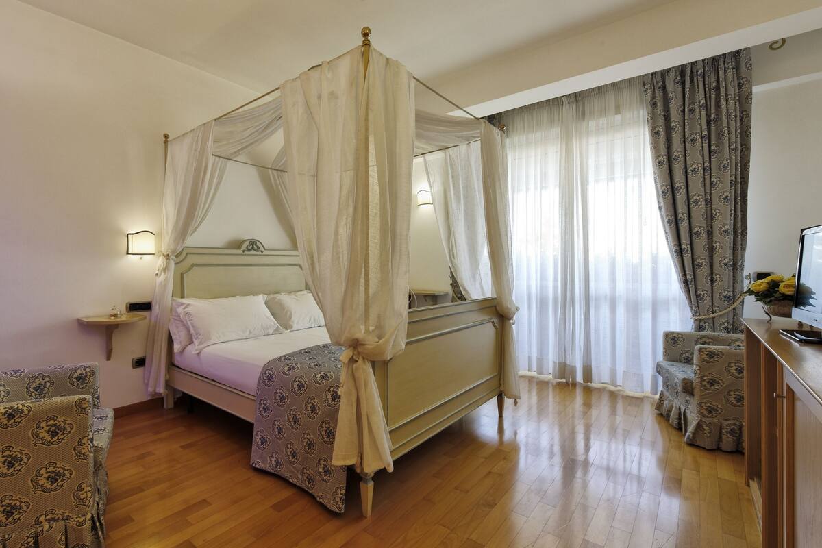 A four-poster canopy bed with a bedside table and lamp, two patterned armchairs, a wooden nightstand, and a large window with sheer curtains.