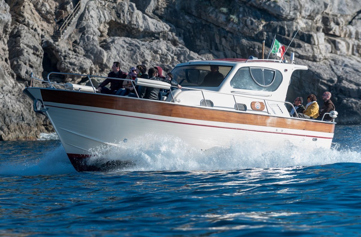 A boat with passengers, moving through water with rocky cliffs in the background.