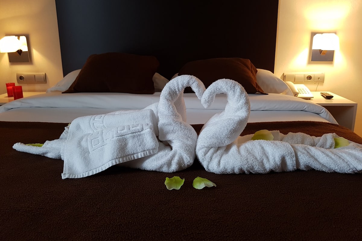 A hotel room with a bed, two wall-mounted lamps, a telephone, and a remote control on the nightstand.