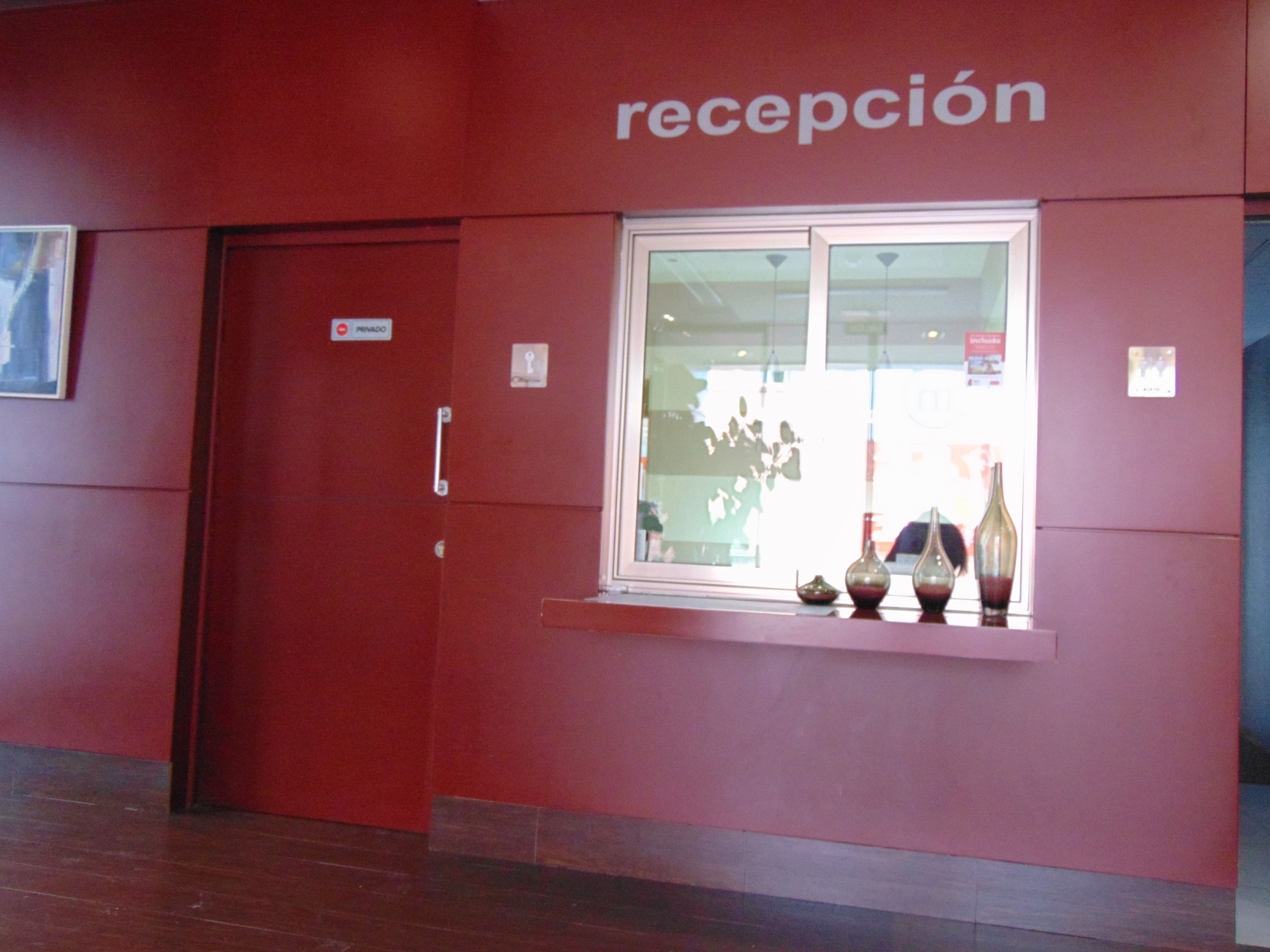 Een receptiebalie met een rode wand, een raam met decoratieve vazen, en een deur met de tekst 'receptie'.