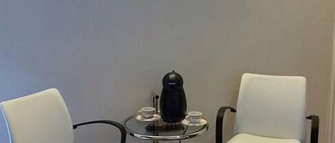 A room with two white chairs, a small round table with a black coffee maker, and a wall-mounted artwork.