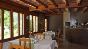 A dining area with wooden beams, large windows, and a stone wall. Tables are set with white tablecloths and chairs. - Hotel Los Nogales (Campo)