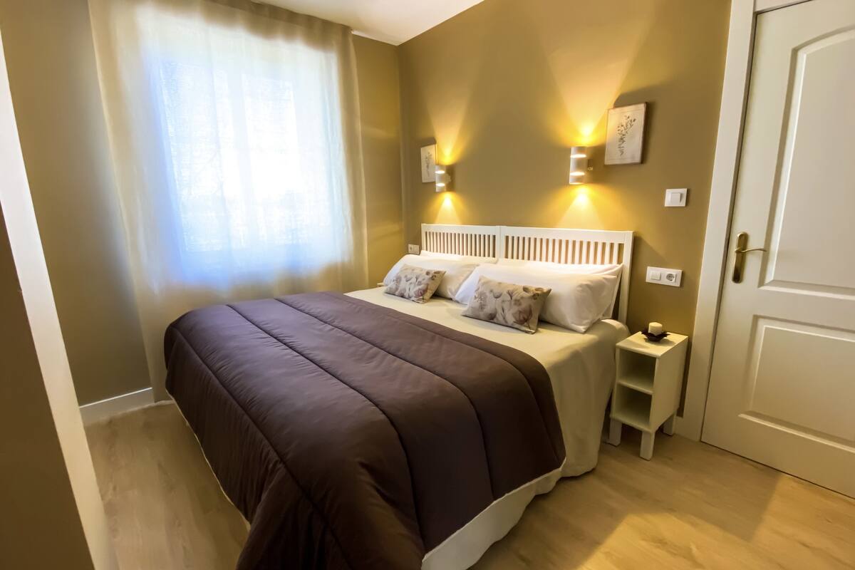 A bedroom with a large bed, two bedside tables, a nightstand, a door, and a window with curtains.