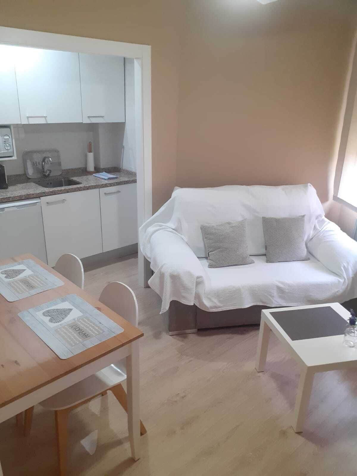 Un espace de vie compact comprenant un coin cuisine, un canapé blanc avec des coussins, une table à manger avec des chaises et une petite table d’appoint.