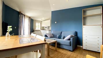 A modern living room with a blue sofa, a dining table with chairs, a kitchen in the background, and a wooden cabinet with drawers.
