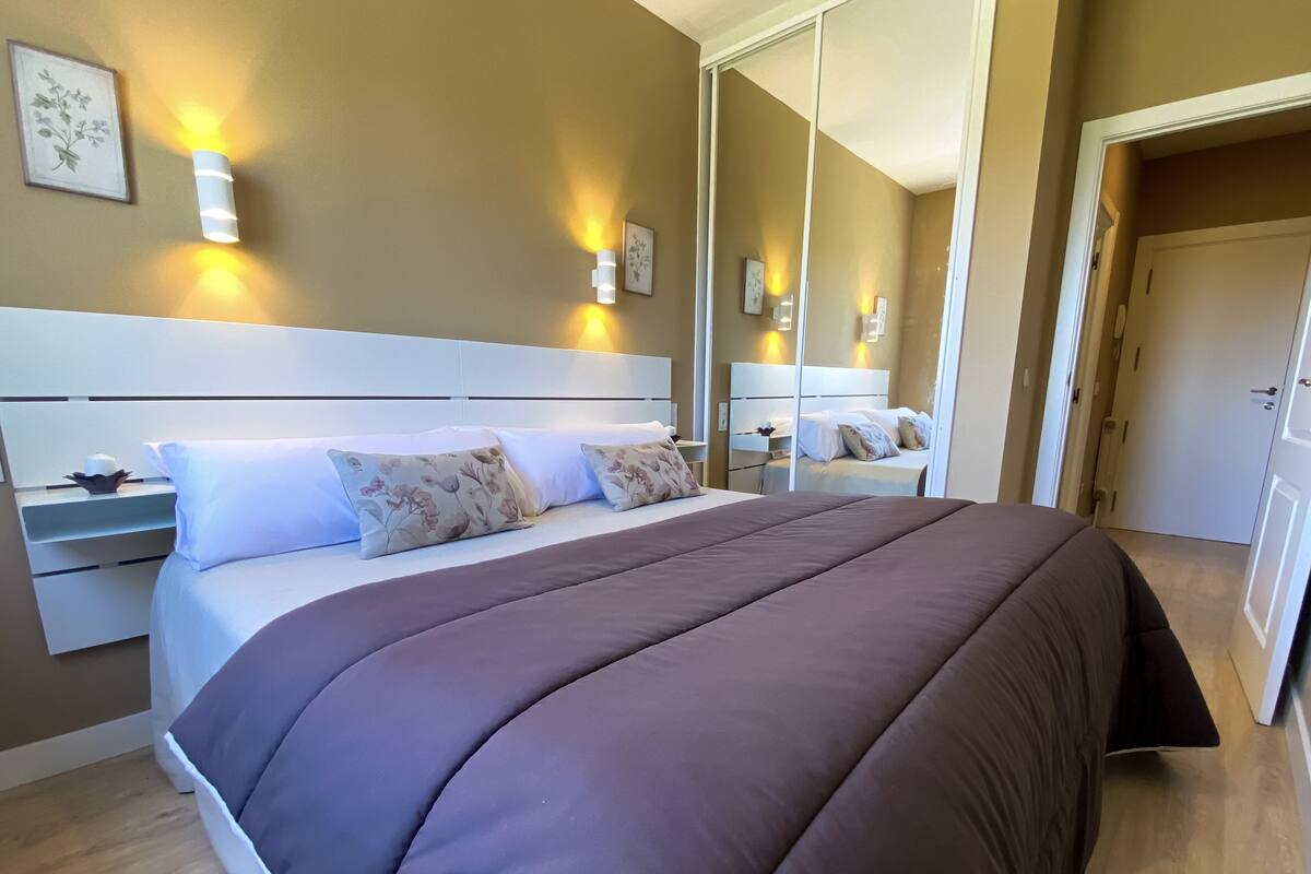A hotel room with a large bed, a bedside table, a mirror, and wall-mounted lights.