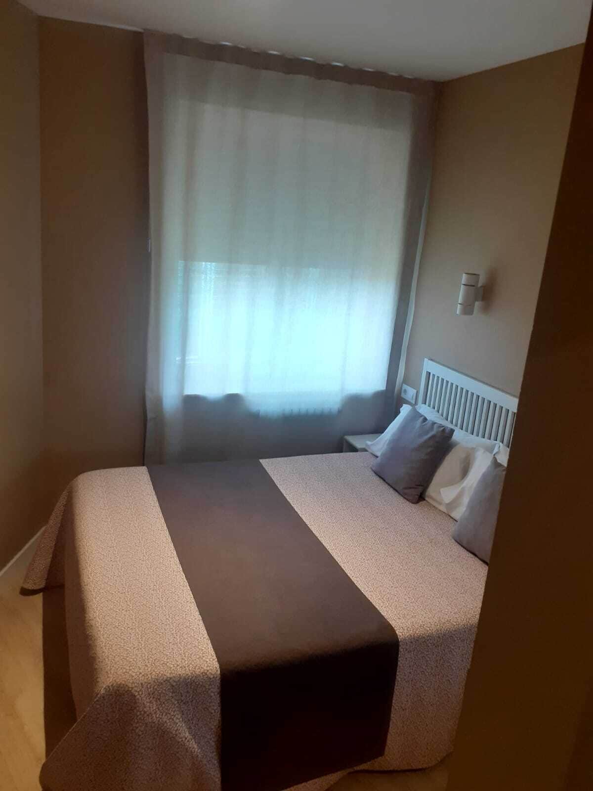 Et hotelværelse med seng, natbord, et vindue med lette gardiner og et termostat på væggen.
