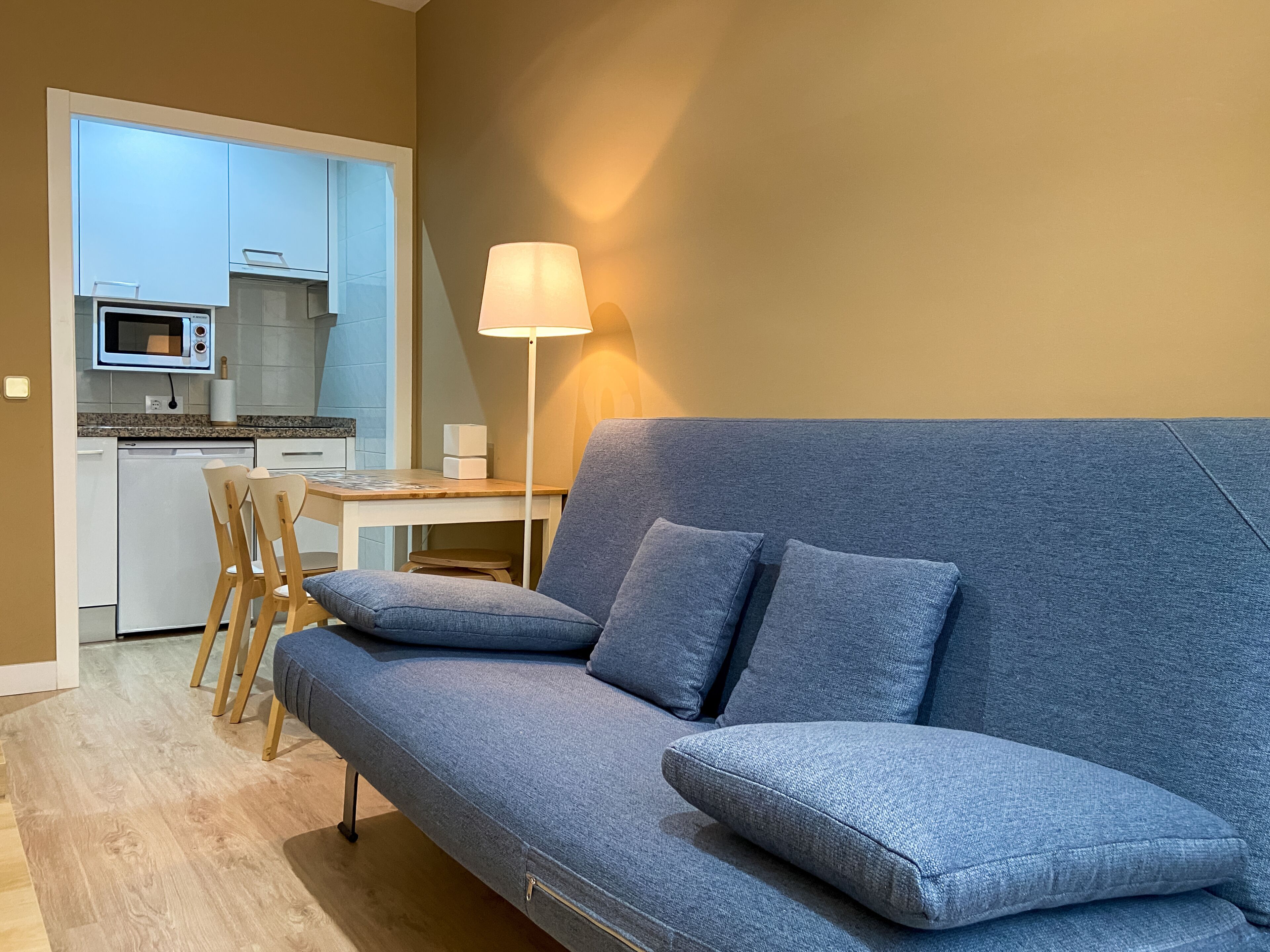 Een moderne woonkamer met een grijze bank, houten stoelen, een eettafel en een keuken op de achtergrond.