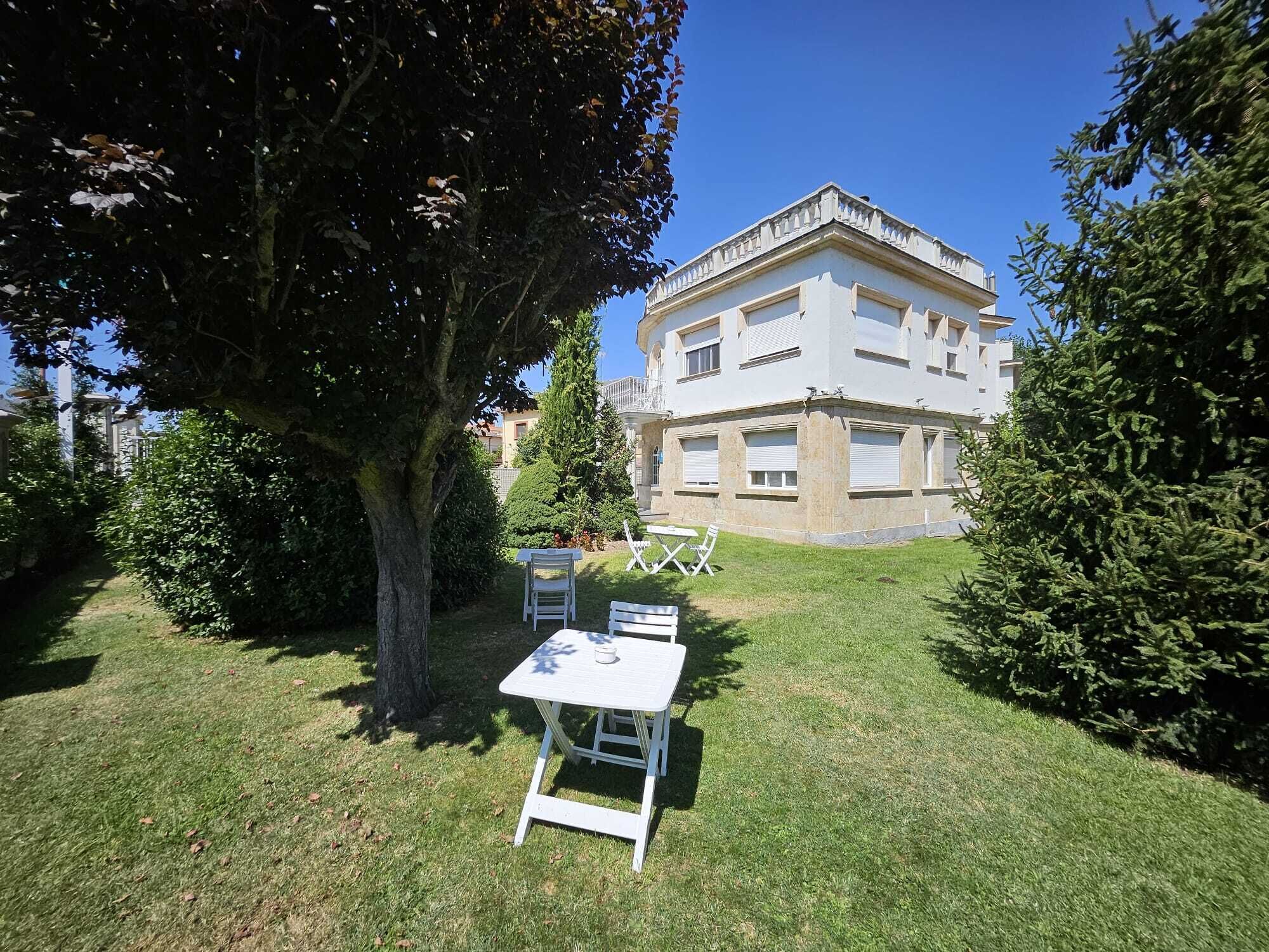 Une cour avec une table et des chaises blanches, entourée de verdure et d’un grand arbre.