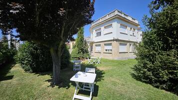 Une cour avec une table et des chaises blanches, entourée de verdure et d’un grand arbre.