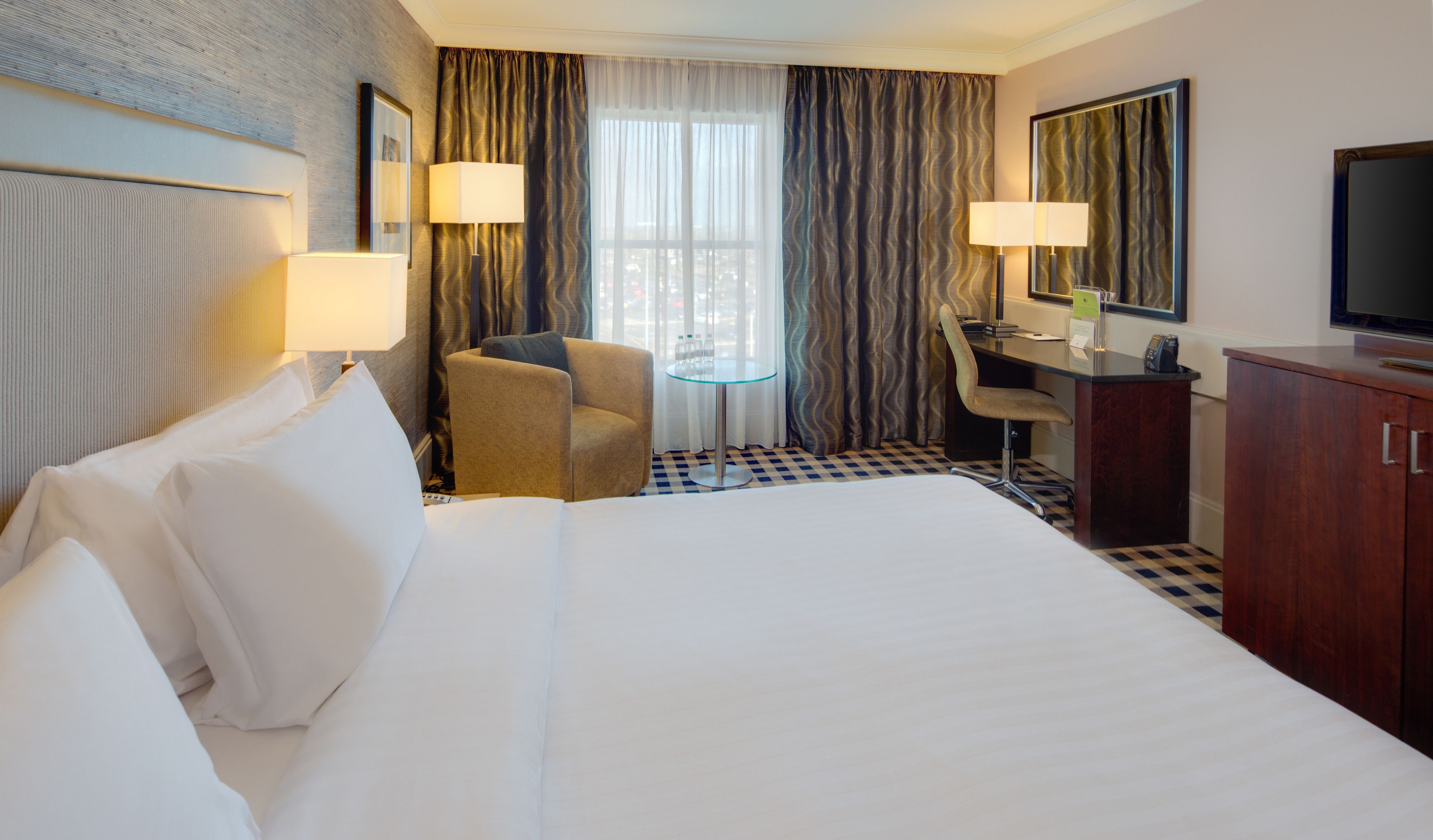 A hotel room with a large bed, a desk with a chair, a small table, and a television.