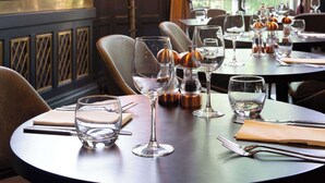 Uma sala de jantar com mesas preparadas para o serviço, um bar com garrafas e uma janela grande com cortinas.