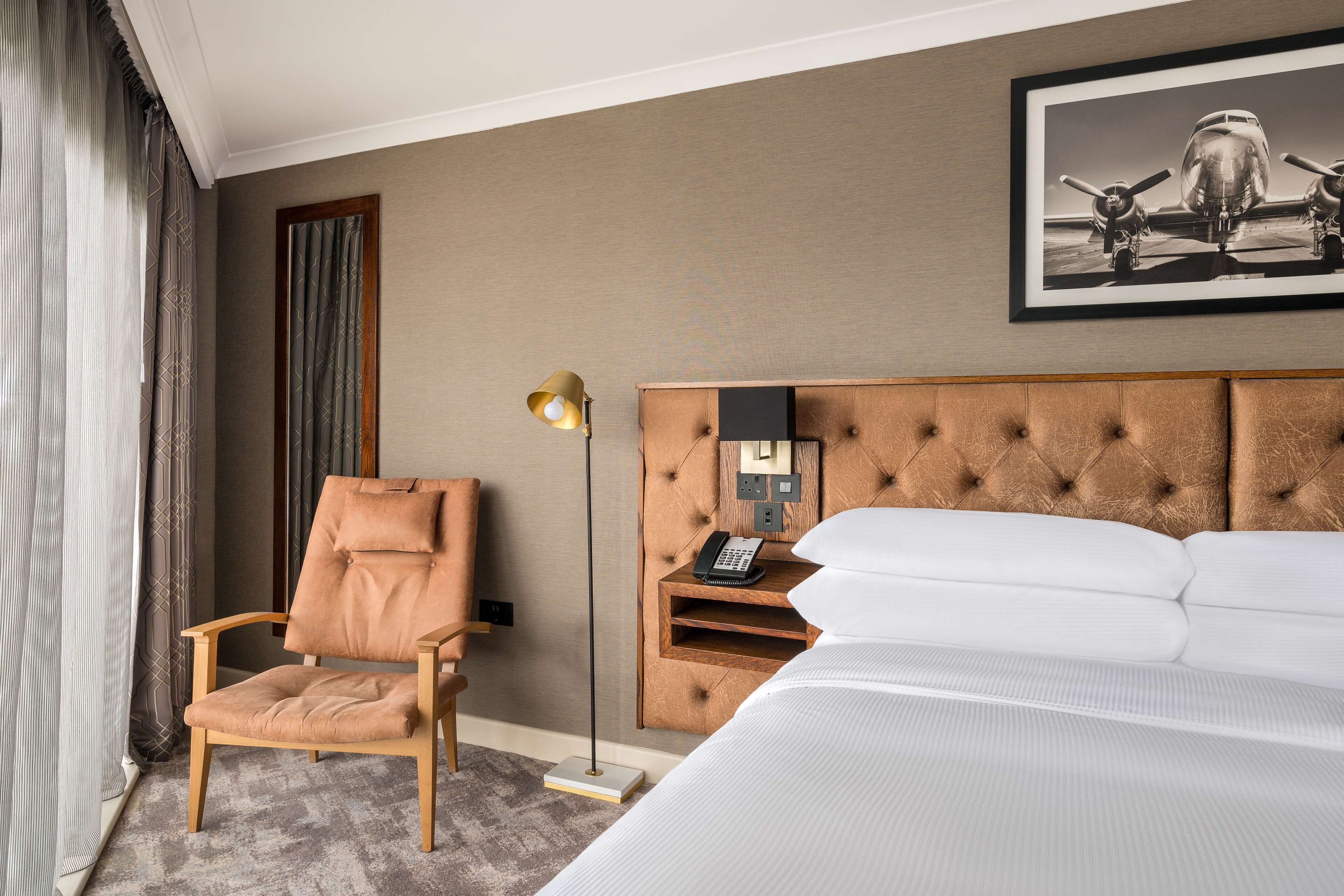 Habitación de hotel con una cama grande, cabecera capitonada, mesita de noche de madera con teléfono, sillón de cuero marrón y lámpara de pie.
