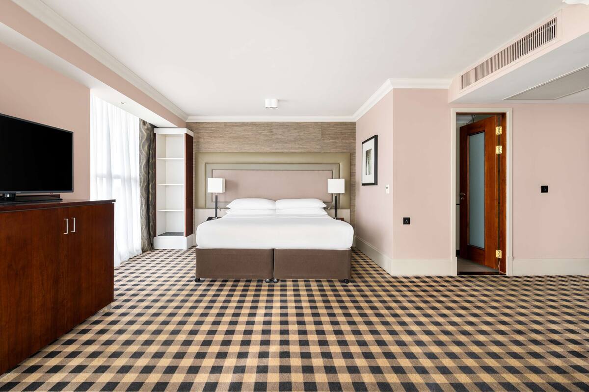 A hotel room with a checkered floor, a bed with two pillows, a TV on a wooden cabinet, and a door leading to another room.