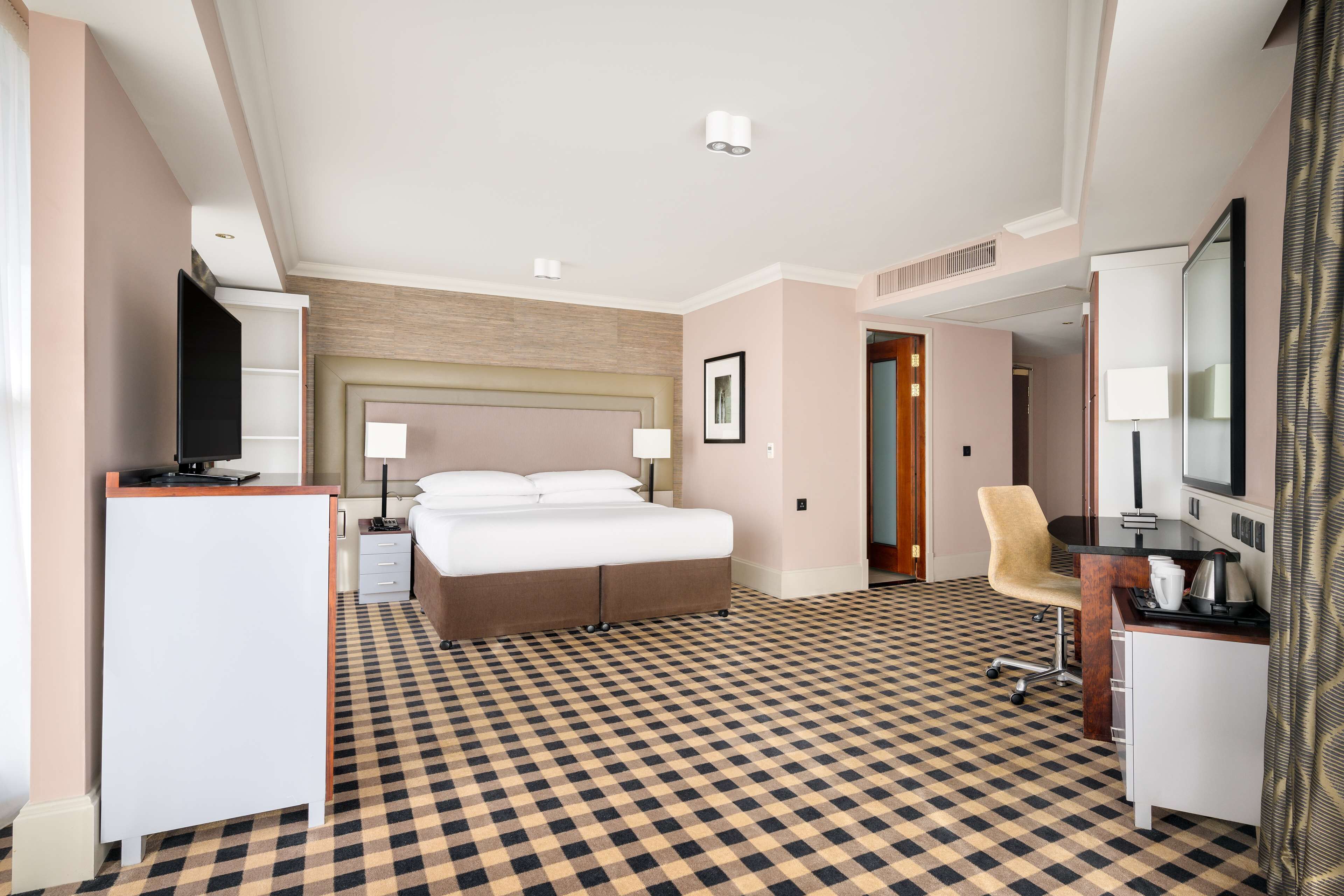 A hotel room with a checkered floor, a bed, a desk, a chair, a TV, and a mirror.