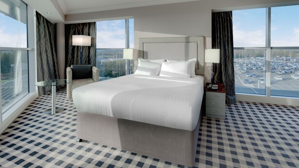 Quarto de hotel com uma cama grande, uma cadeira, uma mesa e vista para um estacionamento através de amplas janelas.