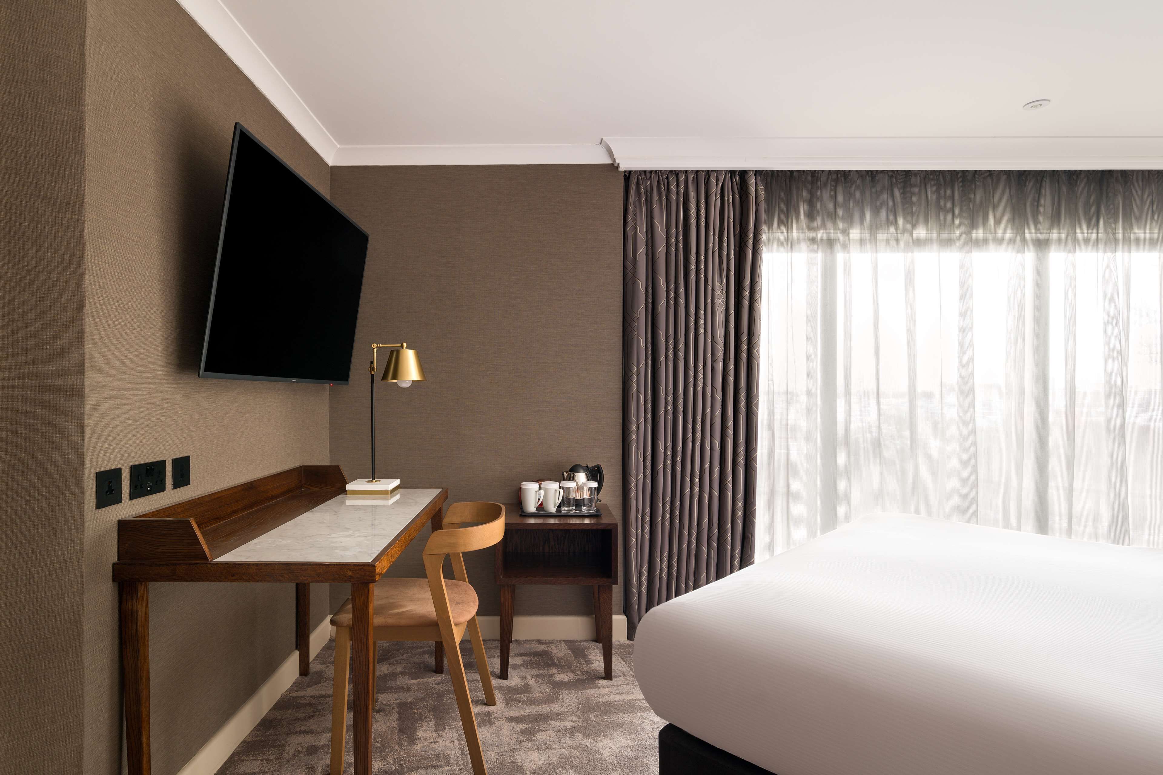 A hotel room with a bed, a desk with a chair, a television mounted on the wall, a lamp, and a window with curtains.