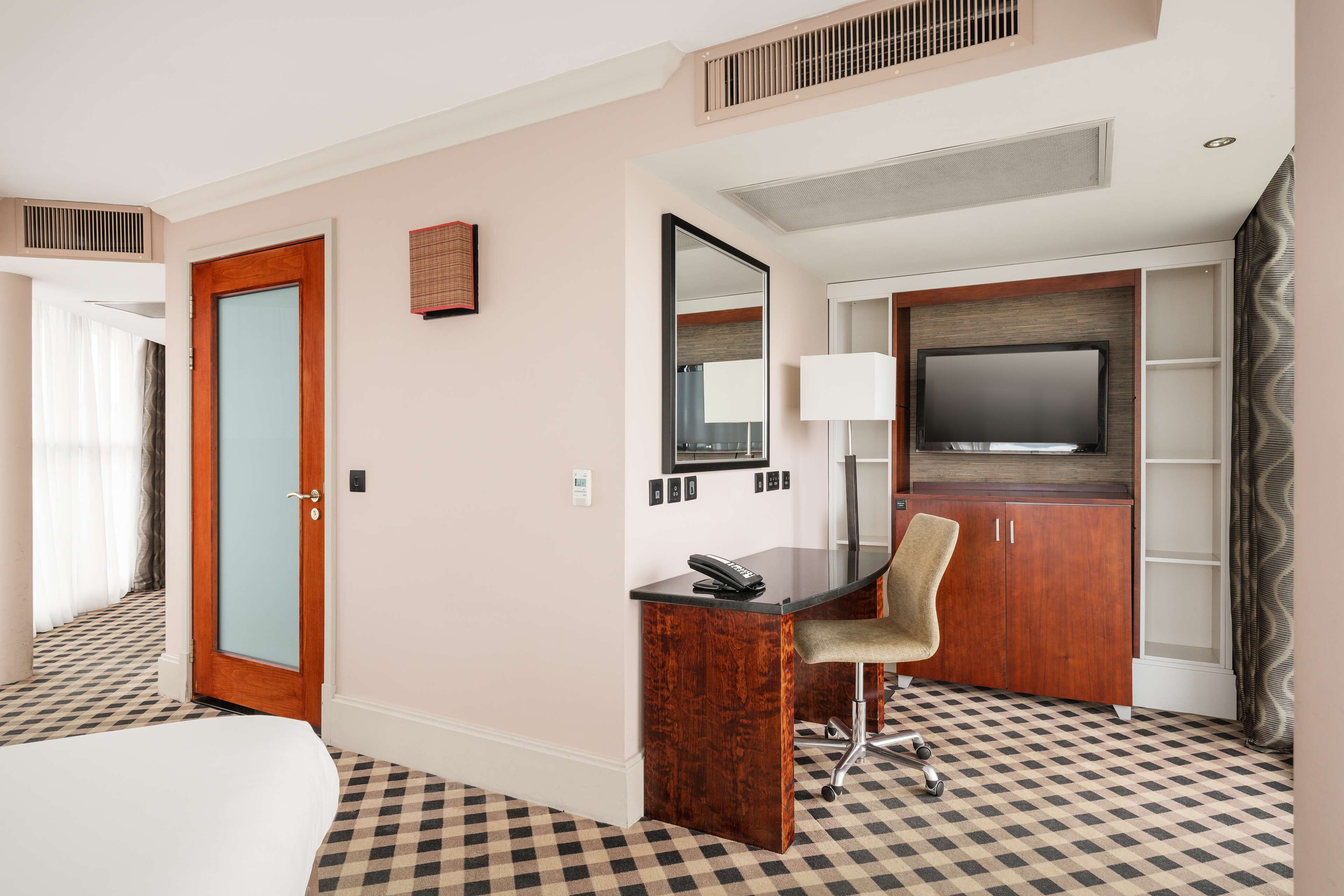 Quarto de hotel com piso xadrez, uma mesa com cadeira, uma televisão e uma porta.