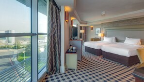 Habitación de hotel con dos camas, un ventanal con vistas a la ciudad, suelo a cuadros y un pequeño escritorio con lámpara.