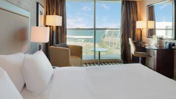A hotel room with a large bed, a desk, a chair, and a view of the cityscape through the window.