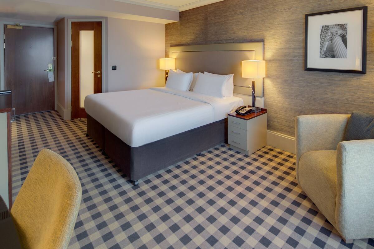 A hotel room with a checkered floor, a bed with white linens, two bedside lamps, a nightstand with a phone, and a framed picture on the wall.
