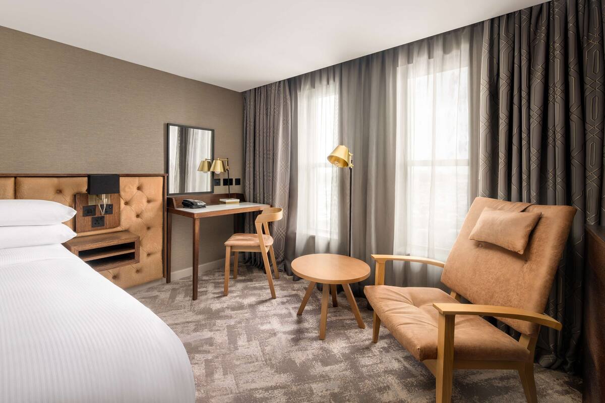 A hotel room with a bed, a desk with a chair, a sofa, and a large window with curtains.