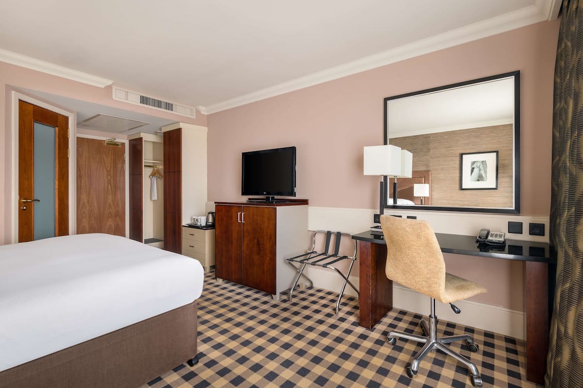 A hotel room with a bed, a desk with a chair, a television, a mirror, and a wardrobe.