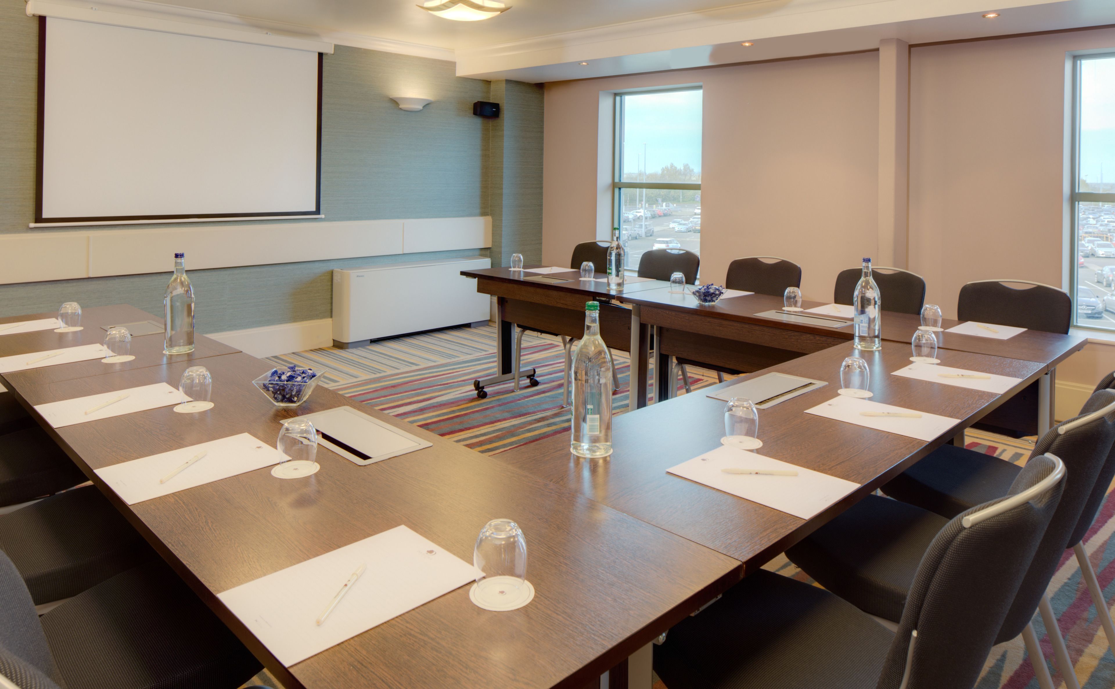A conference room with a long table, chairs, a projector screen, and windows with a view of the city.
