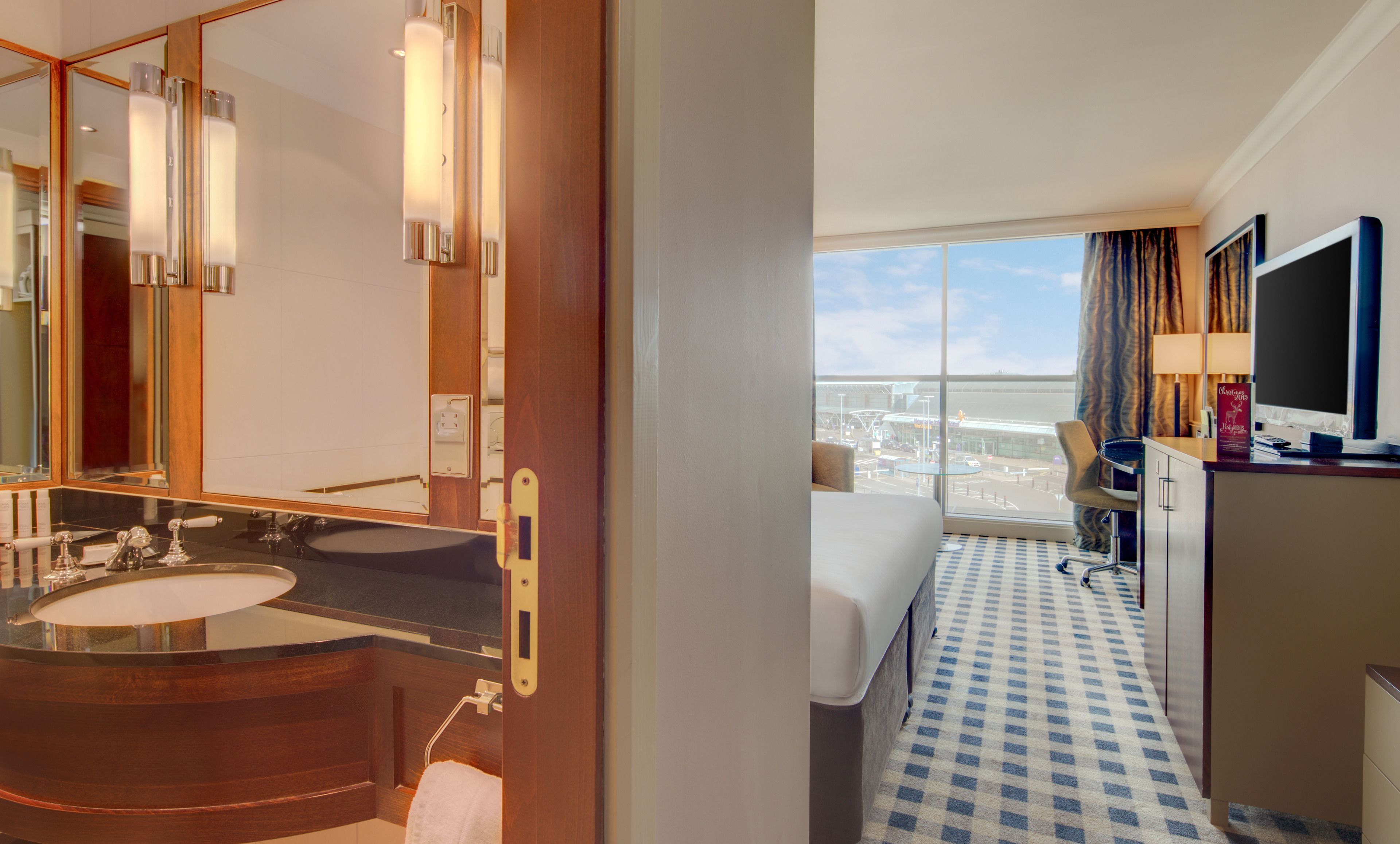 A hotel room with a large window, a bathroom with a double sink, and a bed with a checkered floor.