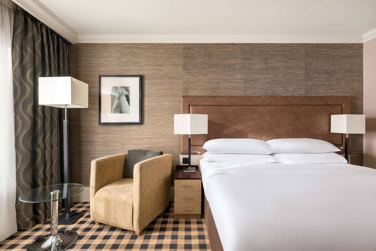 A hotel room with a large bed, two bedside lamps, a chair, a small table, and a framed picture on the wall.