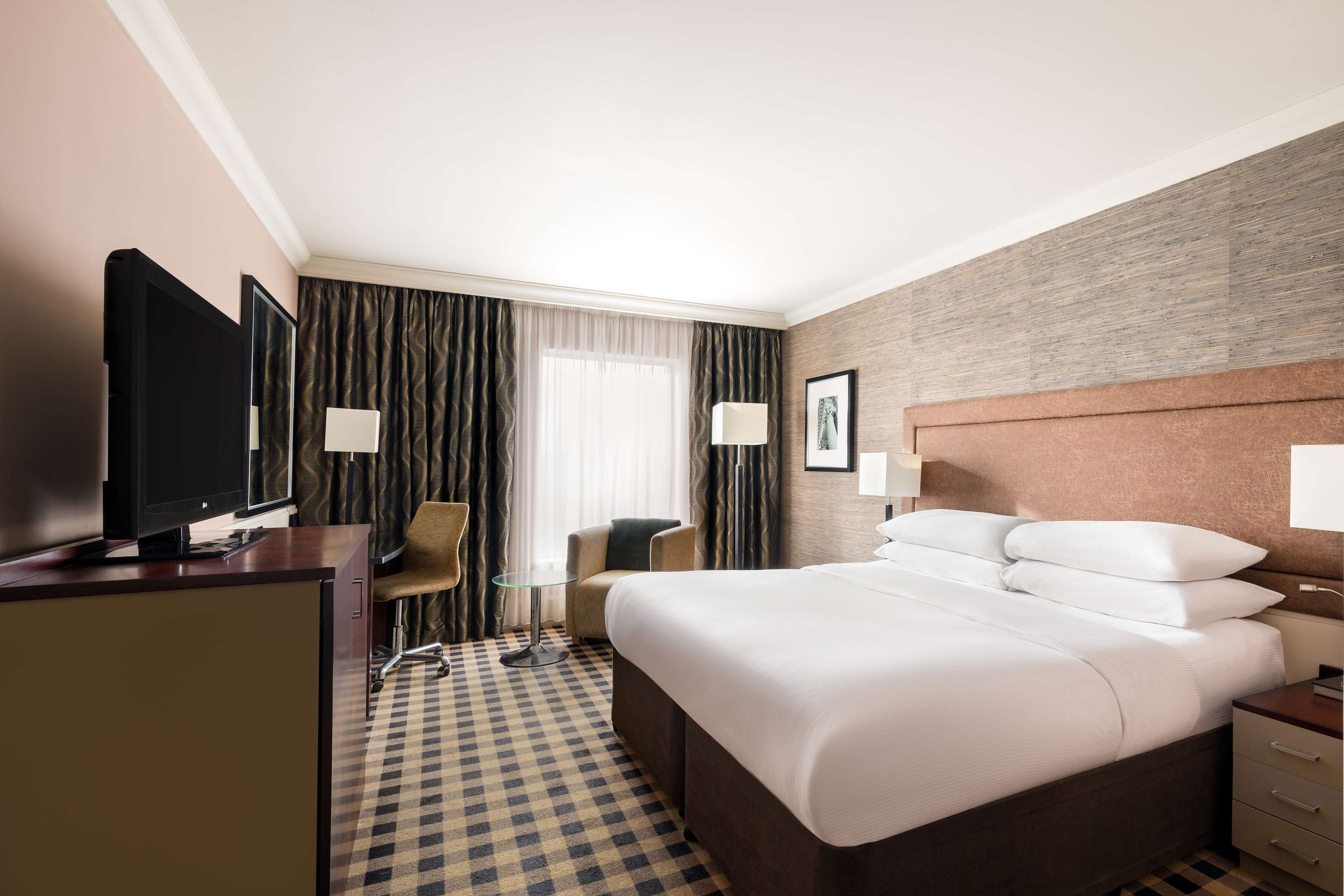 A hotel room with a large bed, a desk with a flat-screen TV, a chair, a small table, and a window with curtains.