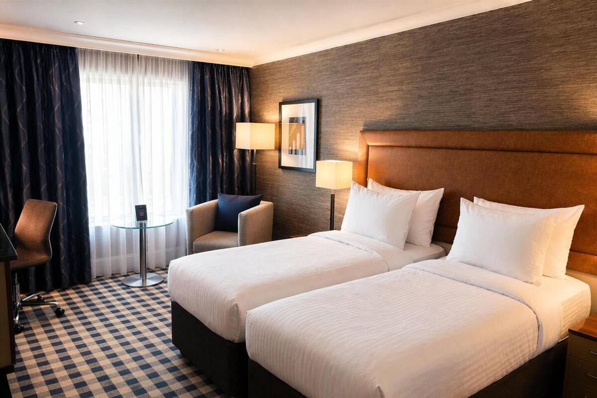 A hotel room with two beds, a desk, a chair, a lamp, and a checkered floor.