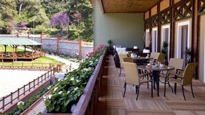 A covered outdoor seating area with tables and chairs, a stone wall, and a view of a landscaped garden and trees. - Padok Premium Hotel and Stables (Ula)