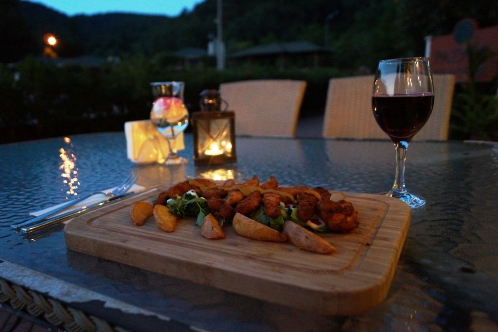 Una mesa con una tabla de madera que contiene comida, una copa de vino tinto y dos velas encendidas.