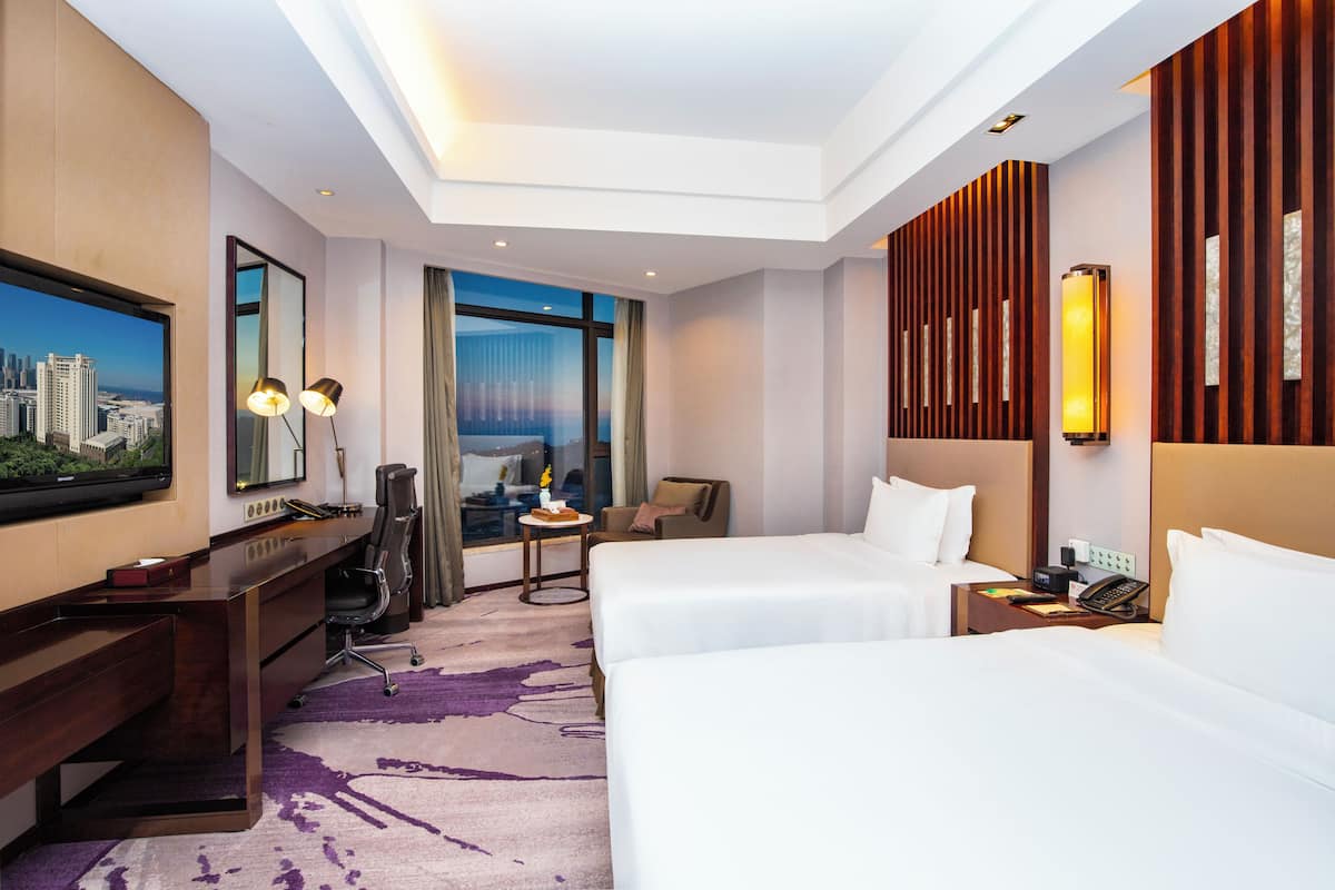 Habitación de hotel con dos camas, un escritorio, una silla, un televisor y un ventanal con vista a la ciudad.