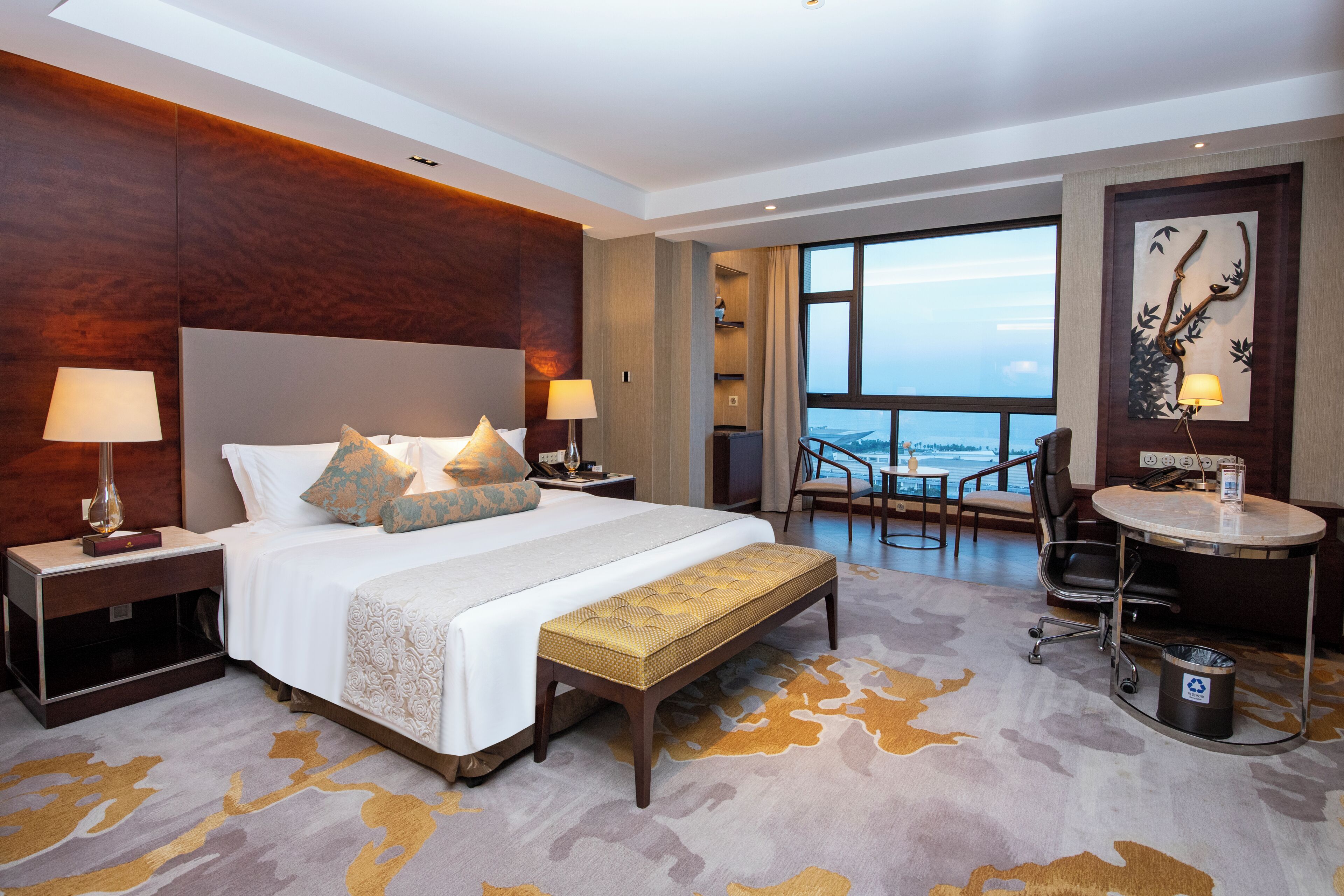 A hotel room with a large bed, two bedside tables, a bench, a desk, a chair, and a view of the city.