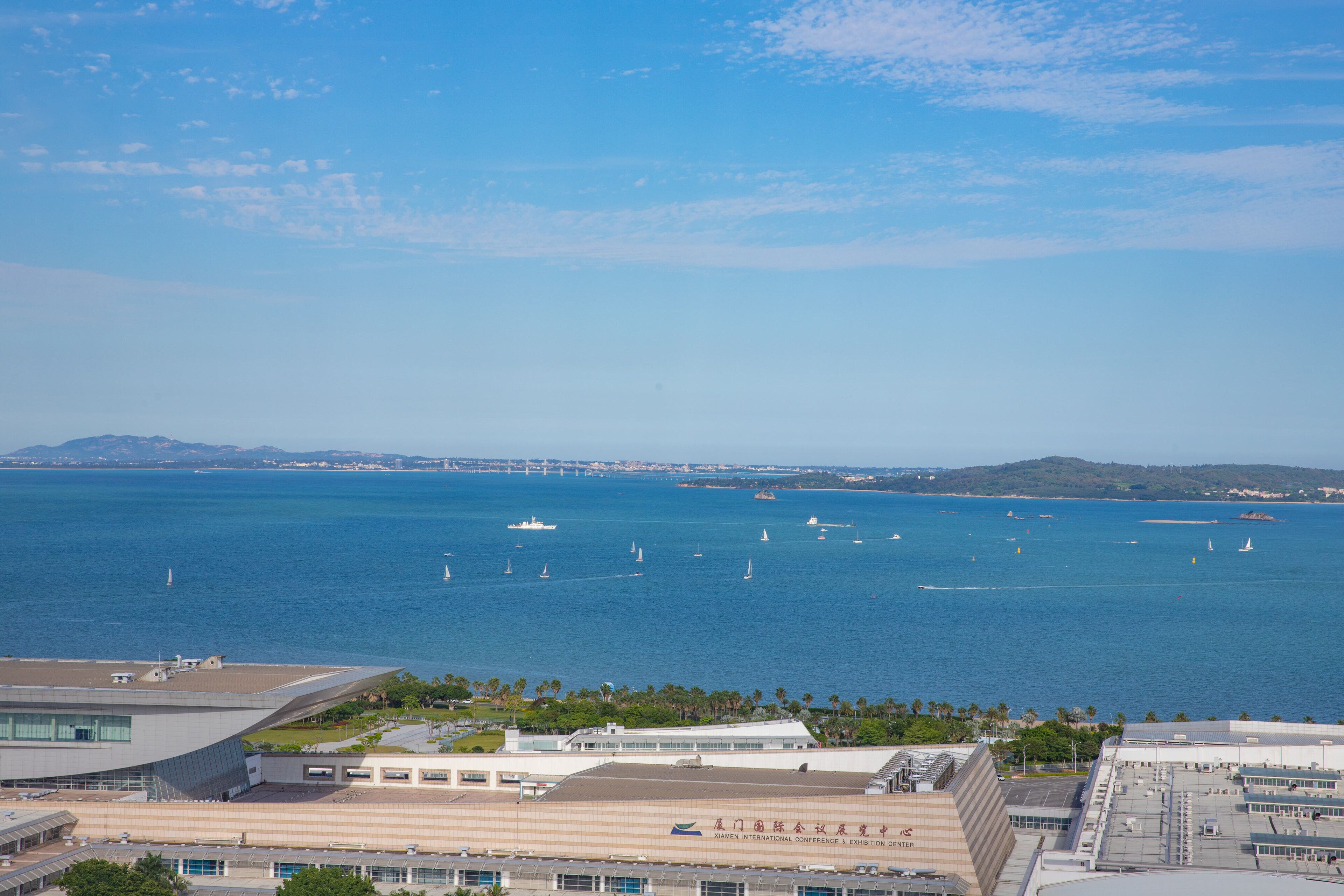 Vista costera con un edificio moderno de gran fachada de vidrio, un puerto deportivo con veleros y un cielo azul despejado.