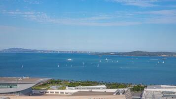 Vista costera con un edificio moderno de gran fachada de vidrio, un puerto deportivo con veleros y un cielo azul despejado.