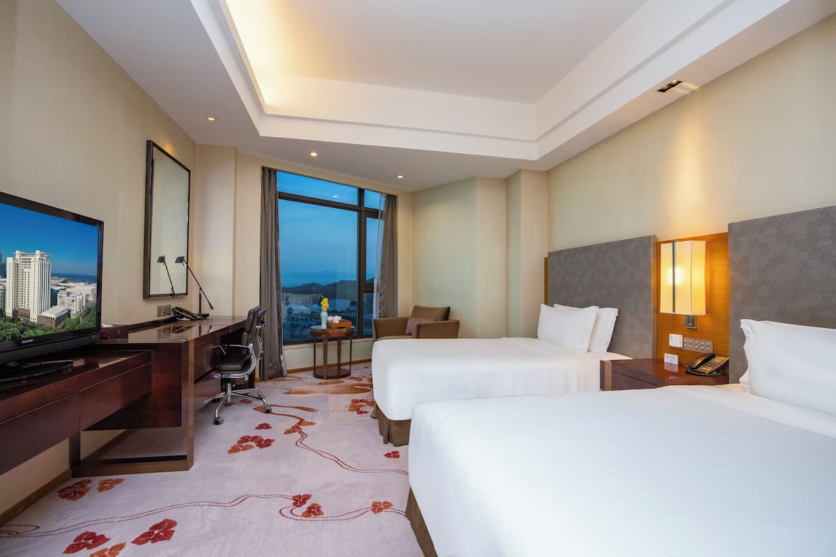 Habitación de hotel con escritorio, dos camas, televisor, ventana grande y vista a la ciudad.
