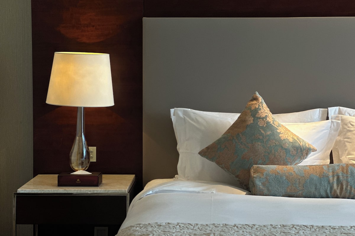 Una cama bien hecha con sábanas blancas y almohadas decorativas, una mesita de noche con lámpara y un cabecero de madera oscura.