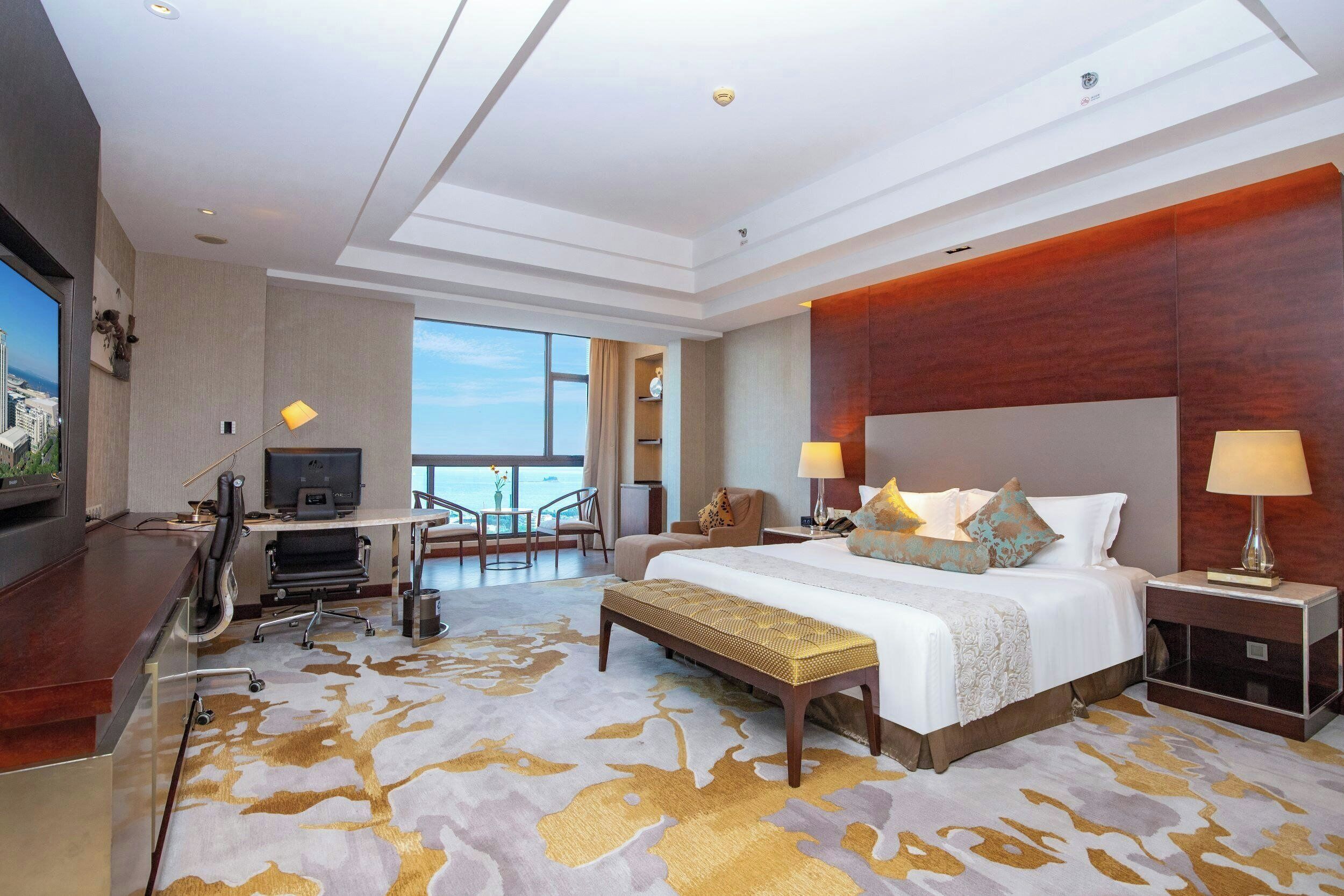 A hotel room with a large bed, a desk with a computer, a TV, and a view of the sea.