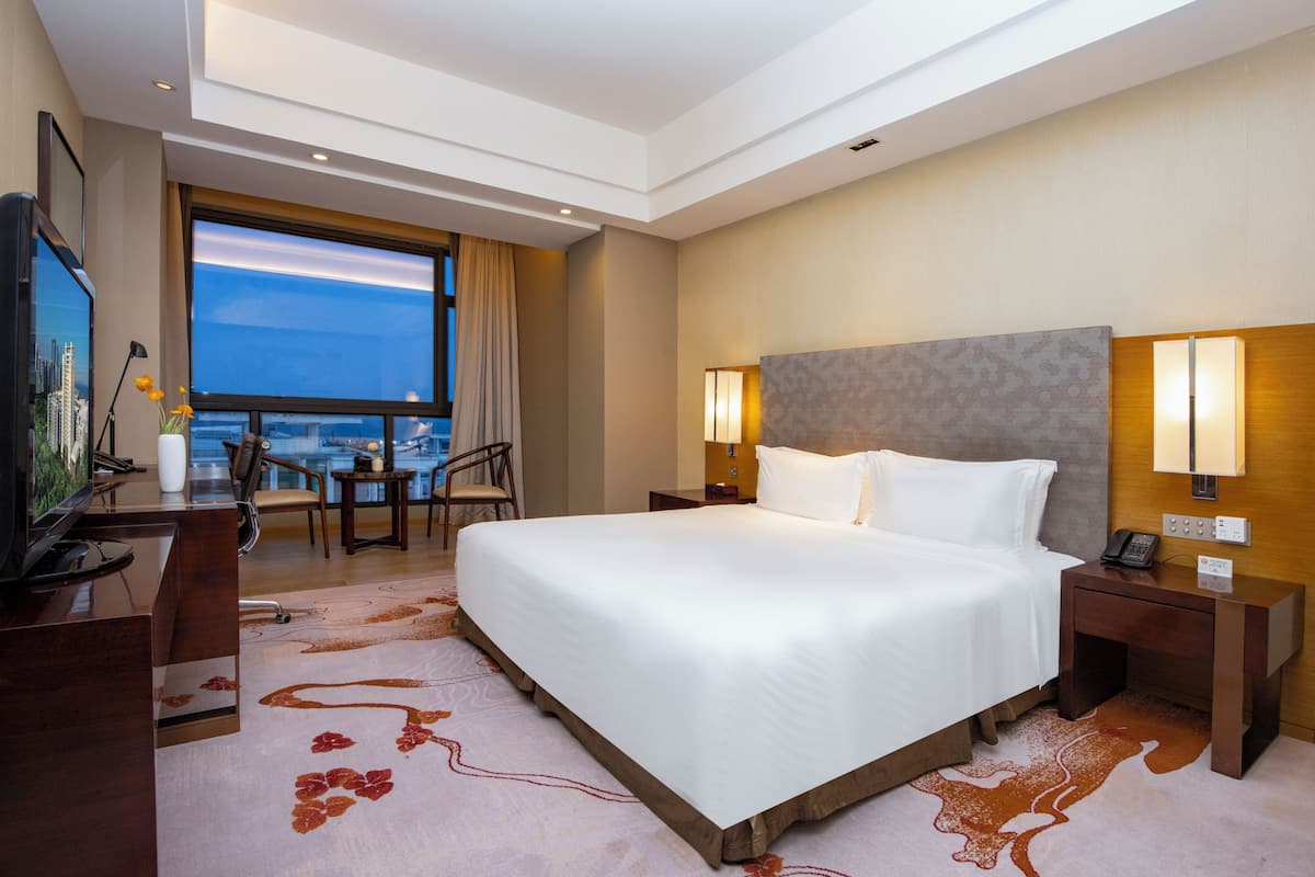 Habitación de hotel con una cama grande, un escritorio con televisor, una lámpara de escritorio, una mesita de noche con teléfono y vistas a la ciudad a través de un ventanal amplio.